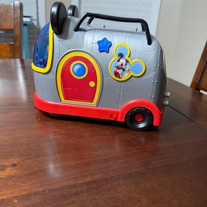 K&ouml;nnte beinhalten: Ein grauer und roter Spielzeugkoffer im Mickey-Mouse-Design mit schwarzen R&auml;dern und einem Griff. Der Koffer hat eine rote T&uuml;r, einen blauen Stern und eine Mickey-Mouse-Grafik. Die Oberseite hat schwarze Mickey-Mouse-Ohren.