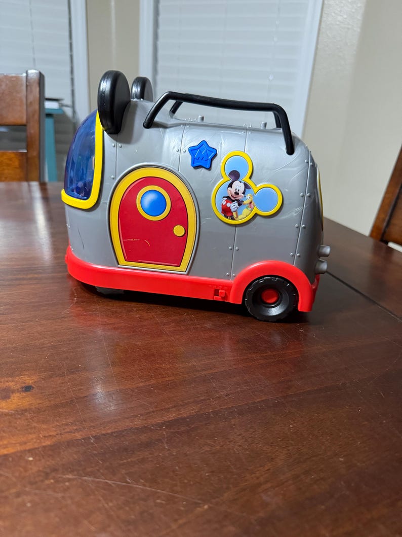 K&ouml;nnte beinhalten: Ein grauer und roter Spielzeugkoffer im Mickey-Mouse-Design mit schwarzen R&auml;dern und einem Griff. Der Koffer hat eine rote T&uuml;r, einen blauen Stern und eine Mickey-Mouse-Grafik. Die Oberseite hat schwarze Mickey-Mouse-Ohren.