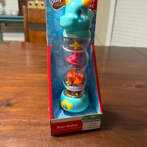 May include: A blue and orange Play Right Rain Maker toy in its original packaging. The toy features a dragon head and a clear tube with colorful elements. The box has the text "12M+ DEVELOPS curiosity, discovery, imagination."