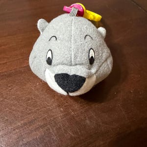 Könnte beinhalten: Ein graues Plüschtier in Form eines Bärenkopfes mit gestickten Augen und einer schwarzen Nase. Es hat kleine Ohren und einen rosa und gelben Kunststoffring oben. Das Spielzeug ist für Babys und Kleinkinder konzipiert.
