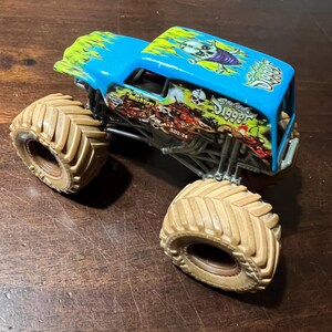 May include: A blue and green toy monster truck with the words "Grave Digger" and a skull graphic. The truck has large tan tires and a detailed chassis. The roof has a flame design.