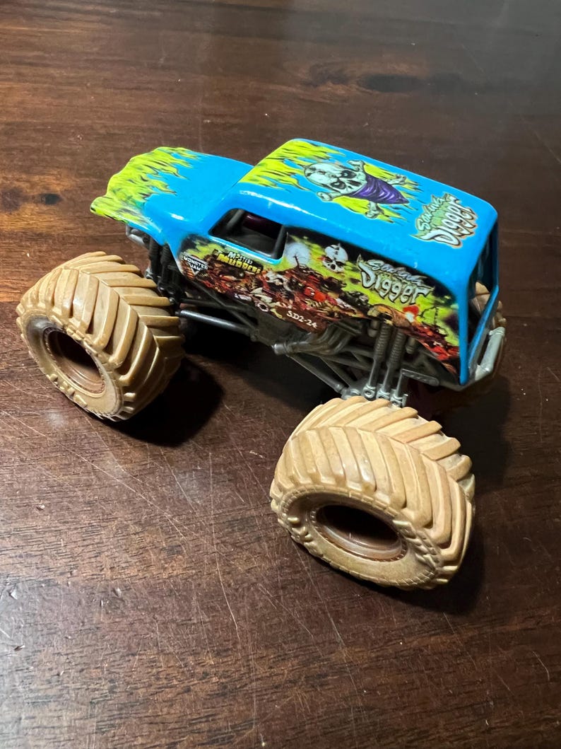 May include: A blue and green toy monster truck with the words "Grave Digger" and a skull graphic. The truck has large tan tires and a detailed chassis. The roof has a flame design.