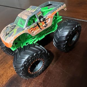 May include: A toy monster truck with a brown and green camouflage design on the cab. The truck has large black tires and a green chassis. The truck has the words "GRAVE DIGGER" and other graphics on the side.