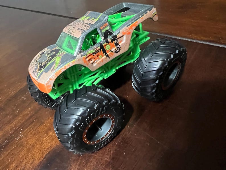 May include: A toy monster truck with a brown and green camouflage design on the cab. The truck has large black tires and a green chassis. The truck has the words "GRAVE DIGGER" and other graphics on the side.