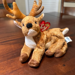 Puede incluir: Un juguete de peluche marrón con forma de reno, con grandes cuernos y una nariz negra. El reno tiene una etiqueta roja en forma de corazón que dice "2000" y "Ty". El juguete está sobre una superficie de madera.
