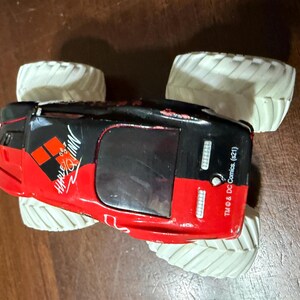 May include: A red, black, and white toy monster truck with large white tires. The truck features the Harley Quinn logo and text. The toy is a licensed DC Comics product.