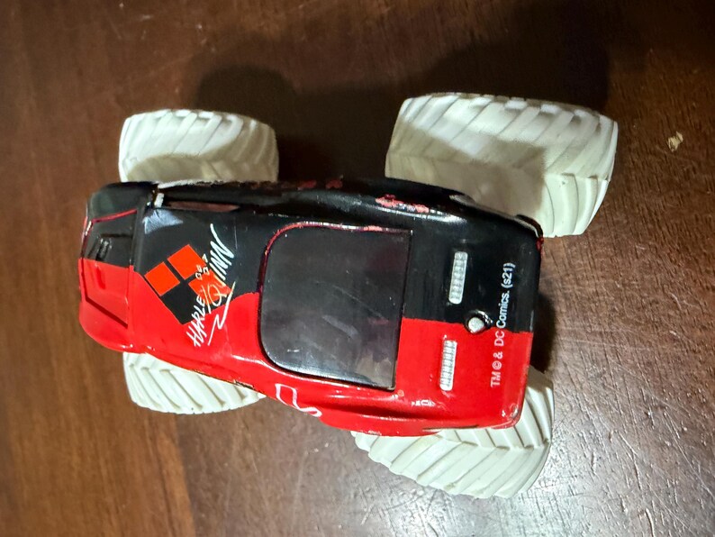 May include: A red, black, and white toy monster truck with large white tires. The truck features the Harley Quinn logo and text. The toy is a licensed DC Comics product.