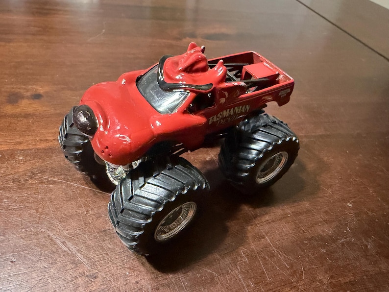 May include: A red toy monster truck with large black tires. The truck has the words "Tasmanian Devil" printed on the side. The truck is sitting on a brown surface.