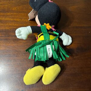 May include: A plush Minnie Mouse doll in a Hawaiian outfit. The doll has a black head with pink bow, white gloves, and yellow shoes. It wears a green grass skirt and a yellow and black top. The tag reads "Hula Minnie".