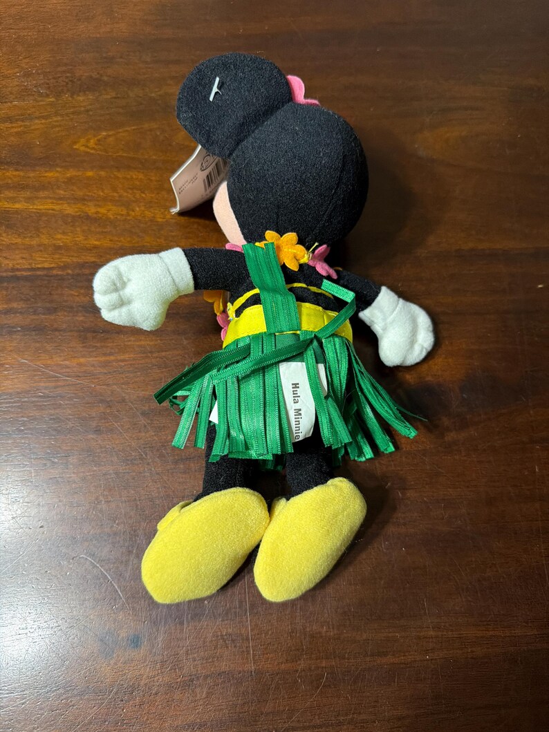 May include: A plush Minnie Mouse doll in a Hawaiian outfit. The doll has a black head with pink bow, white gloves, and yellow shoes. It wears a green grass skirt and a yellow and black top. The tag reads "Hula Minnie".