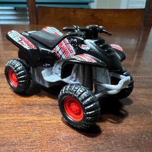 Puede incluir: Un quad de juguete negro con gráficos rojos y blancos. El quad tiene cuatro ruedas y un asiento.