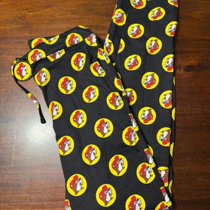 May include: Black pajama pants with a repeating pattern of a cartoon beaver head inside a yellow circle. The beaver wears a red hat and has a white face. The pants have a drawstring and are made of a soft material.