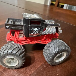 May include: A black and red toy monster truck with large, black tires. The truck features a skull and crossbones design on the side and the name "BONE SHAKER". The truck has a detailed engine and suspension.