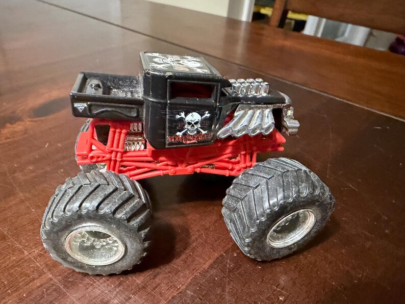 May include: A black and red toy monster truck with large, black tires. The truck features a skull and crossbones design on the side and the name "BONE SHAKER". The truck has a detailed engine and suspension.