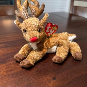 Puede incluir: Un juguete de peluche de reno con pelaje marrón, detalles blancos y una nariz roja. El reno tiene grandes cuernos y una etiqueta que dice "2000". El juguete está tumbado sobre una superficie de madera.