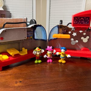 K&ouml;nnte beinhalten: Ein rotes, graues und gelbes Spielzeug-Spielset mit Mickey Mouse, Minnie Mouse und Donald Duck Figuren. Das Spielset hat eine rote Basis, graue W&auml;nde und einen roten Deckel mit einem Bildschirm. Die Figuren tragen bunte Outfits.