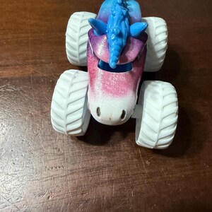 May include: A toy monster truck with a unicorn design. The truck has a pink, purple, and blue body with a blue mane and white tyres. The toy is designed to look like a unicorn.