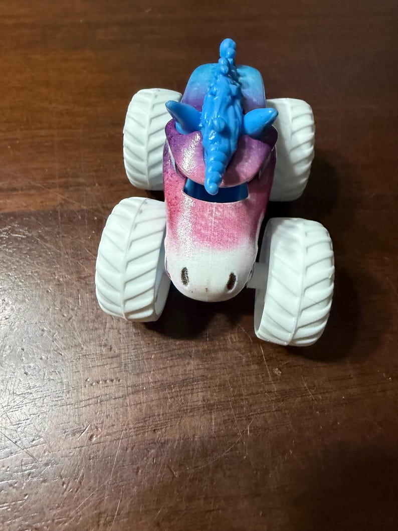 May include: A toy monster truck with a unicorn design. The truck has a pink, purple, and blue body with a blue mane and white tyres. The toy is designed to look like a unicorn.