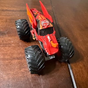May include: Red and orange toy monster truck with large black tires. The truck has a detailed design with a logo on the hood. The toy is on a wooden surface.