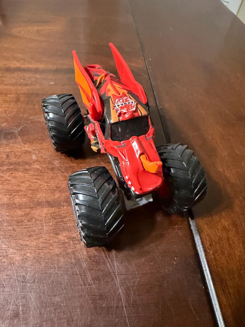 May include: Red and orange toy monster truck with large black tires. The truck has a detailed design with a logo on the hood. The toy is on a wooden surface.
