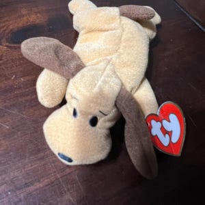Puede incluir: Un juguete de peluche con forma de perro en color beige con orejas y detalles marrones. El perro tiene ojos negros y una nariz negra. Una etiqueta roja en forma de corazón con texto blanco está adjunta.