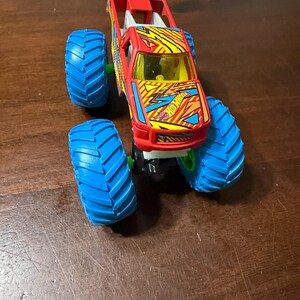 Puede incluir: Un juguete de cami&oacute;n monstruo Hot Wheels rojo y amarillo con neum&aacute;ticos azules. El cami&oacute;n tiene un dise&ntilde;o detallado con el logotipo de Hot Wheels y un parabrisas amarillo. Los grandes neum&aacute;ticos azules tienen un dibujo profundo.