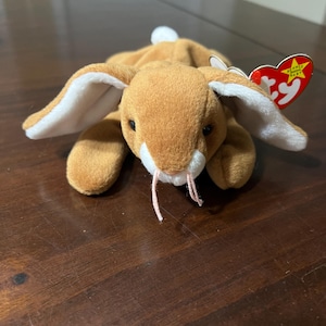 May include: A plush brown rabbit toy with long ears, white inner ear lining, and a small white tail. The toy has black eyes and pink whiskers. A red heart-shaped tag is attached to the ear.