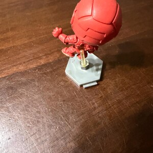 May include: A red figurine of a character in a dynamic pose, mounted on a gray hexagonal base. The figure has a spherical head and detailed suit design. The base is supported by a clear plastic stand.