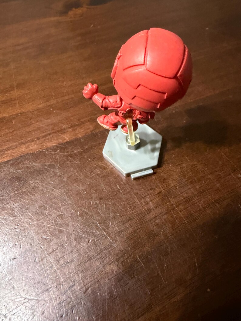 May include: A red figurine of a character in a dynamic pose, mounted on a gray hexagonal base. The figure has a spherical head and detailed suit design. The base is supported by a clear plastic stand.