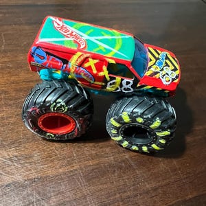 May include: A Hot Wheels monster truck toy in red, green, and blue. It has black tyres with yellow detailing and the number "88" on the side. The roof displays the Hot Wheels logo.