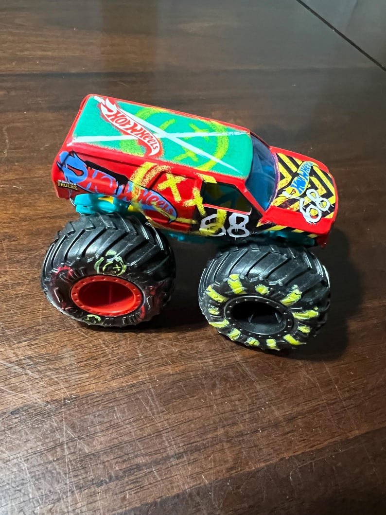 May include: A Hot Wheels monster truck toy in red, green, and blue. It has black tyres with yellow detailing and the number "88" on the side. The roof displays the Hot Wheels logo.