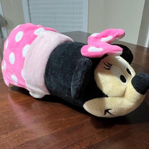 May include: A plush Minnie Mouse toy lying down. The toy has a black body, a light pink bow with white polka dots, and a pink and white polka dot wrap. The face is cream-coloured with black eyes and a black nose.
