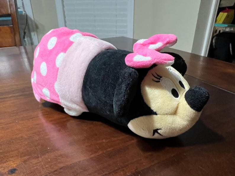 May include: A plush Minnie Mouse toy lying down. The toy has a black body, a light pink bow with white polka dots, and a pink and white polka dot wrap. The face is cream-coloured with black eyes and a black nose.