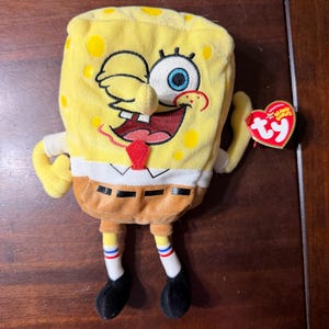 May include: A plush Spongebob Squarepants toy with a yellow rectangular body, brown pants, and white socks. The character has a winking expression, a red tie, and a heart-shaped red Ty Beanie Babies tag attached to the left arm.