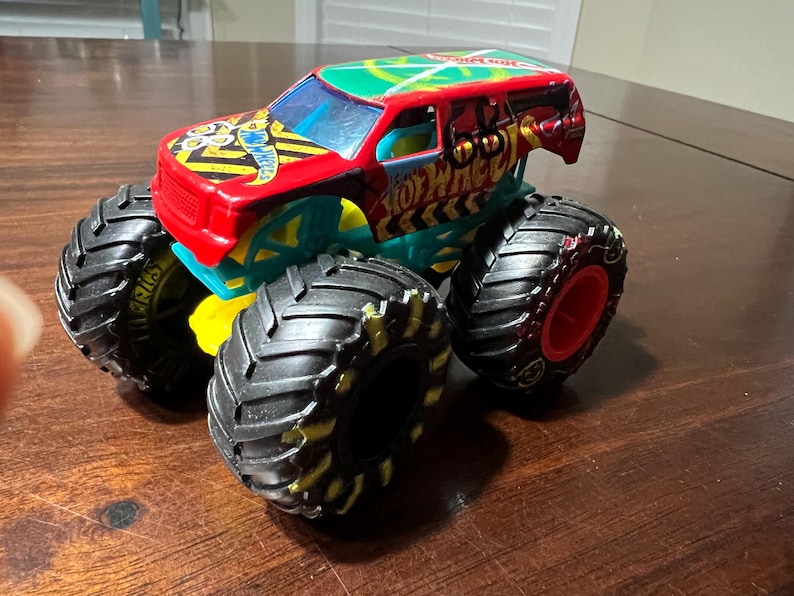 May include: A brightly coloured Hot Wheels monster truck toy. The truck has a red, green, and teal body with black tyres and yellow accents. The tyres are large with a detailed tread pattern. The truck has the Hot Wheels logo.