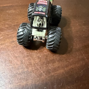 May include: Black and green Grave Digger monster truck toy with large black tires. The truck has the words "Grave Digger" and "BAD BONE" printed on the top. The truck has a clear plastic windshield.