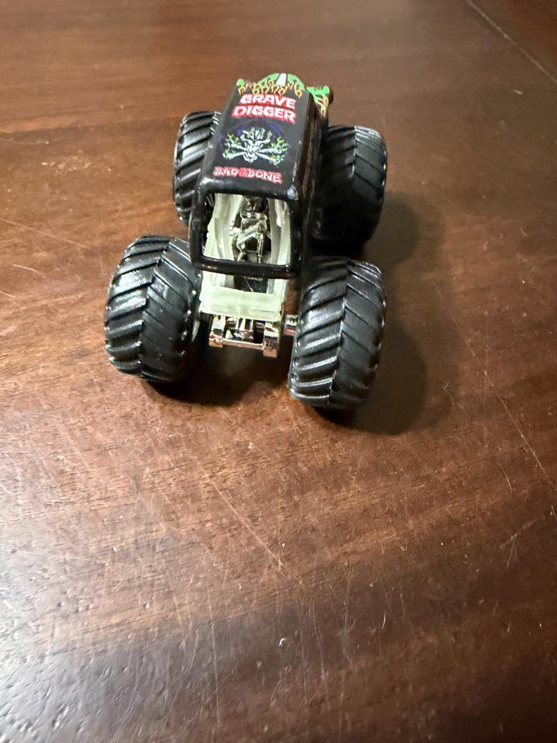 May include: Black and green Grave Digger monster truck toy with large black tires. The truck has the words "Grave Digger" and "BAD BONE" printed on the top. The truck has a clear plastic windshield.