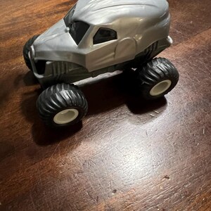 May include: A grey monster truck toy with black tyres and white wheels. The truck has a unique design with a rounded, almost helmet-like, cab.