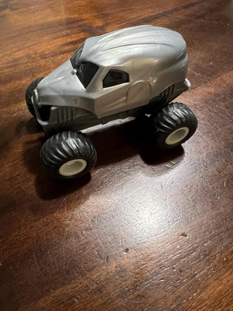 May include: A grey monster truck toy with black tyres and white wheels. The truck has a unique design with a rounded, almost helmet-like, cab.