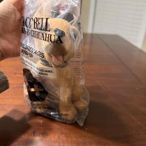 May include: A Taco Bell plush toy chihuahua in a clear plastic bag. The plush dog is tan with black ears and a black nose. The bag has text that reads "Taco Bell" and "Children Ages and Up."