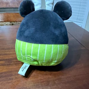 May include: A plush toy with a black head, large round ears, and a green and white striped base. The ears are white with pink inner details. A Disney tag is attached.