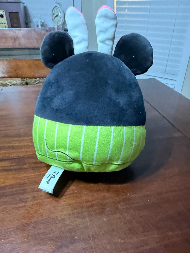May include: A plush toy with a black head, large round ears, and a green and white striped base. The ears are white with pink inner details. A Disney tag is attached.