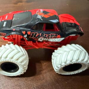 May include: A red and black toy monster truck with the words "Harley Quinn" and heart graphics. The truck has large white tires and a black interior.