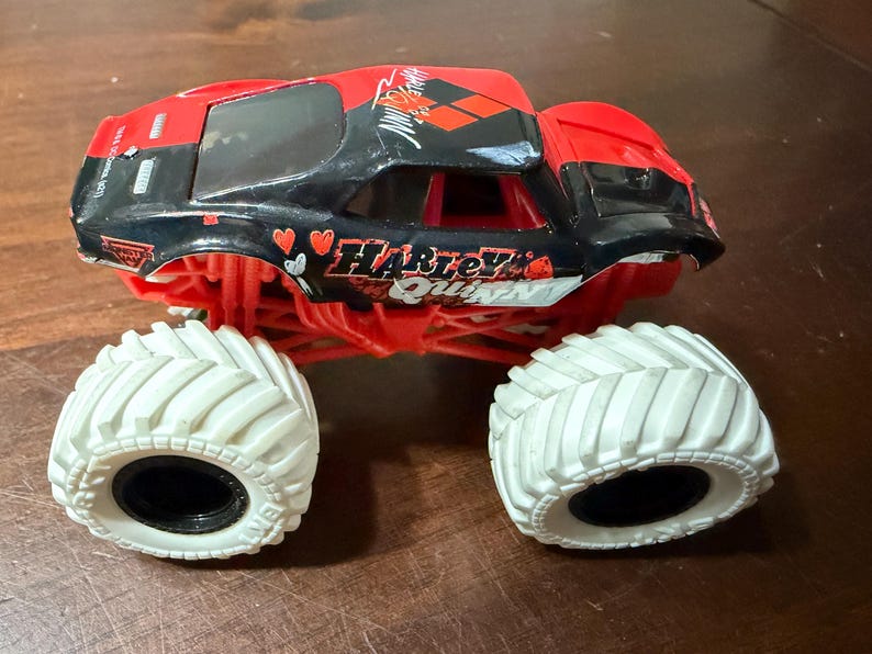 May include: A red and black toy monster truck with the words "Harley Quinn" and heart graphics. The truck has large white tires and a black interior.