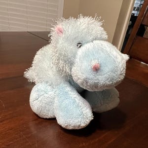 May include: A plush, light blue hippo stuffed animal with a pink nose and ears. The toy is sitting on a dark brown wooden surface. The hippo has a soft, fuzzy texture.