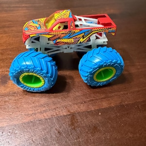 Puede incluir: Un juguete de cami&oacute;n monstruo Hot Wheels rojo, amarillo y azul con neum&aacute;ticos azules grandes y un chasis blanco. El cami&oacute;n presenta el logotipo de Hot Wheels y un dise&ntilde;o detallado. Los neum&aacute;ticos tienen un interior verde.