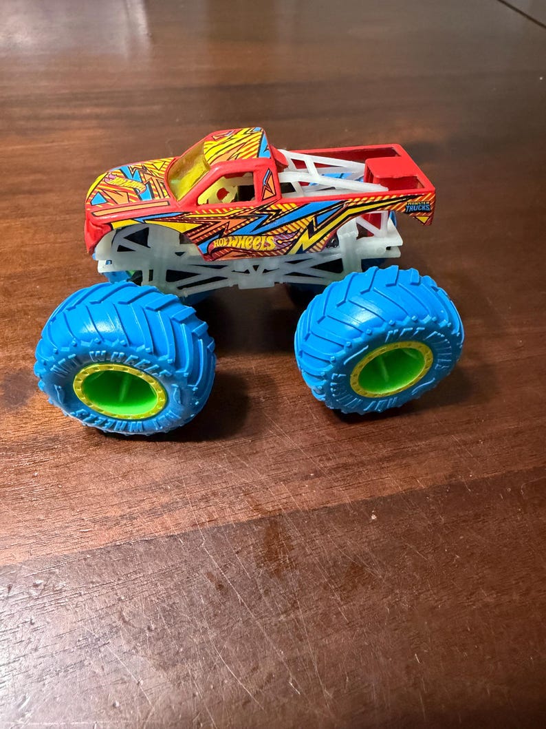Puede incluir: Un juguete de cami&oacute;n monstruo Hot Wheels rojo, amarillo y azul con neum&aacute;ticos azules grandes y un chasis blanco. El cami&oacute;n presenta el logotipo de Hot Wheels y un dise&ntilde;o detallado. Los neum&aacute;ticos tienen un interior verde.