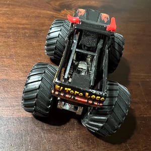Op de afbeelding: Zwarte en oranje speelgoed monstertruck met grote zwarte banden. De truck heeft rode hoorns en de woorden "El Toro Loco" zijn zichtbaar aan de zijkant. De truck staat op een bruin oppervlak.