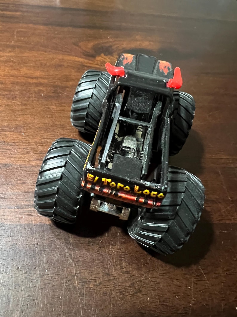 Op de afbeelding: Zwarte en oranje speelgoed monstertruck met grote zwarte banden. De truck heeft rode hoorns en de woorden "El Toro Loco" zijn zichtbaar aan de zijkant. De truck staat op een bruin oppervlak.