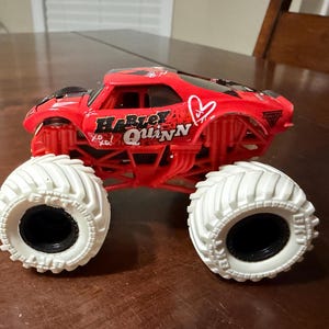 May include: A red monster truck toy with white tires. The truck features the name "Harley Quinn" in white and black lettering, along with a heart and "xo xo" accents. The tires have black centers.